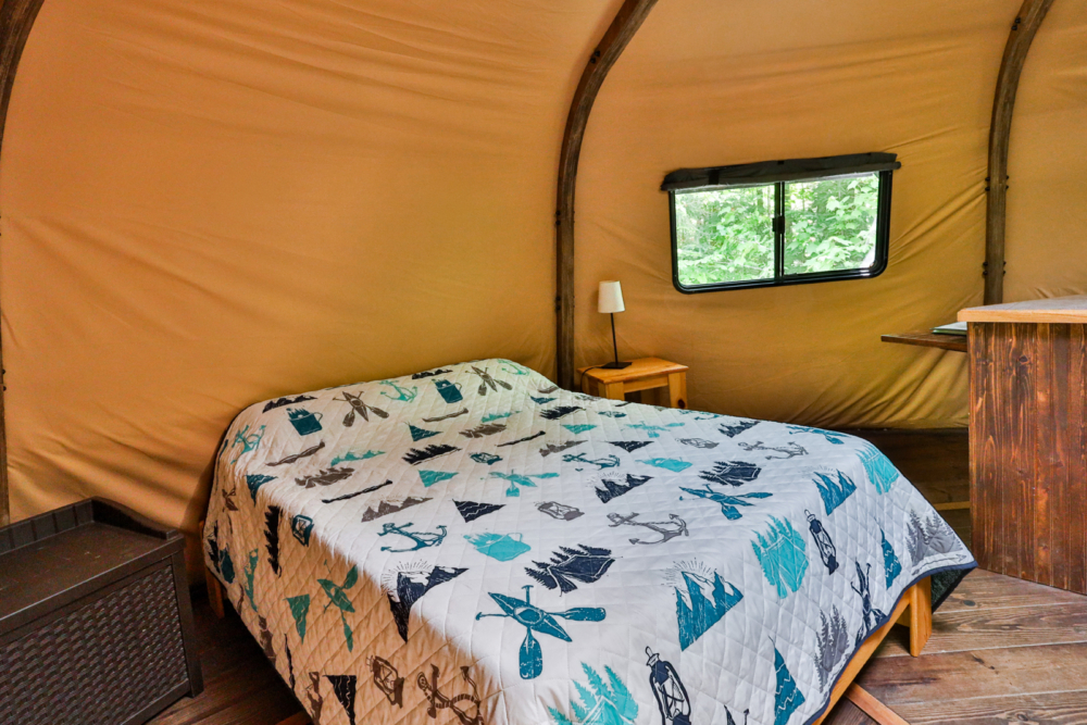Intérieur douillet d’une yourte style glamping avec lit double, literie à motifs et fenêtre donnant sur la forêt.