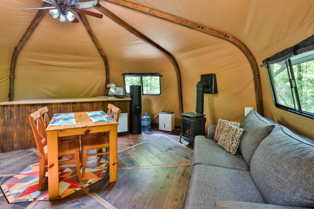 Intérieur chaleureux d’une yourte avec table à manger, sofa, poêle à bois et fenêtres donnant sur la forêt.