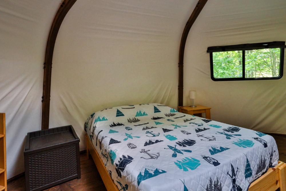 Intérieur d’une yourte avec lit double, literie à motifs de plein air et fenêtre donnant sur la forêt.