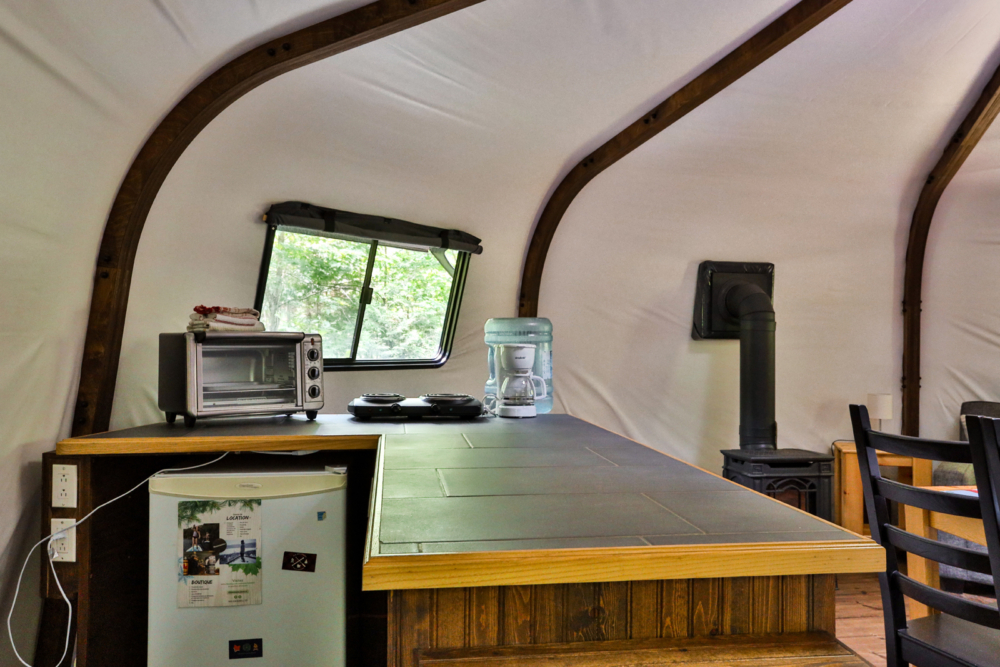 Intérieur d’une yourte avec coin cuisine, comptoir en bois, réfrigérateur, électroménagers et poêle à bois.