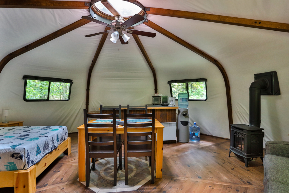 Intérieur chaleureux d’une yourte avec lit, table à manger, cuisinière à bois et ventilateur au plafond.