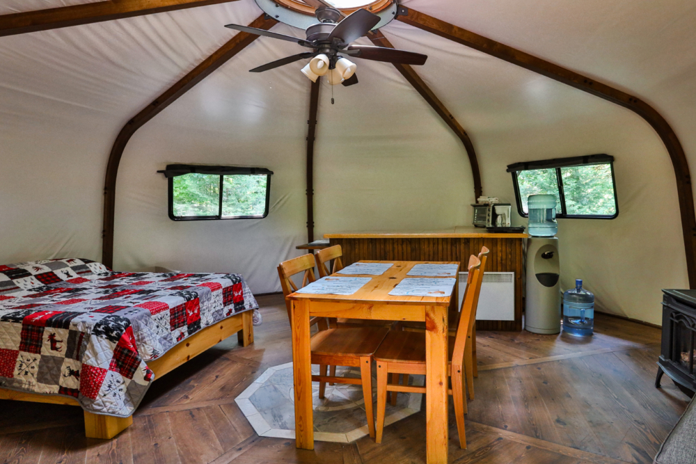 Intérieur d’une yourte avec lit, table à manger en bois, ventilateur au plafond et poêle à bois.