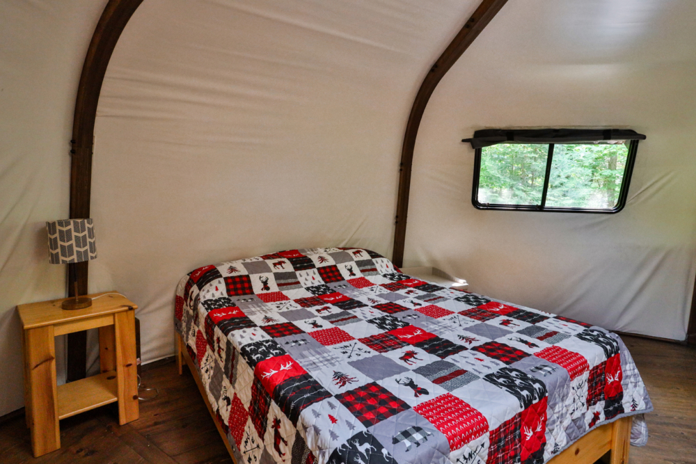 Intérieur d’une yourte avec lit double, courtepointe à carreaux et fenêtre donnant sur la forêt.