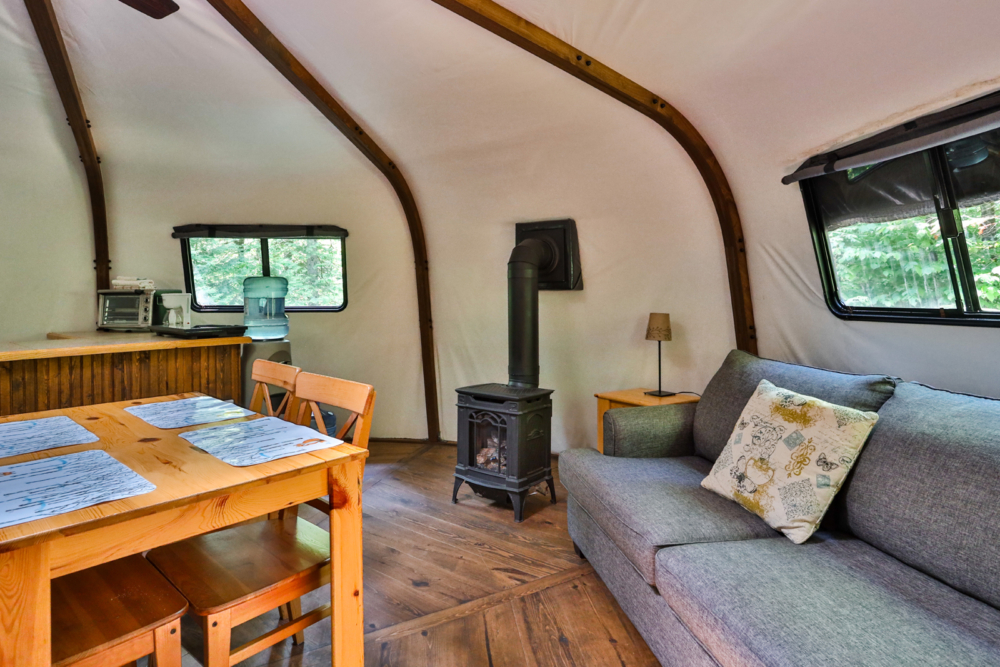 Intérieur chaleureux d’une yourte avec poêle à bois, canapé, table à manger et fenêtres donnant sur la nature.