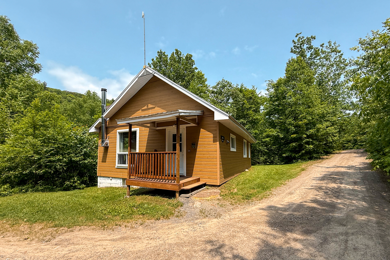 Chalet avec petit balcon couvert, entouré d’arbres et accessible par un chemin de gravier.