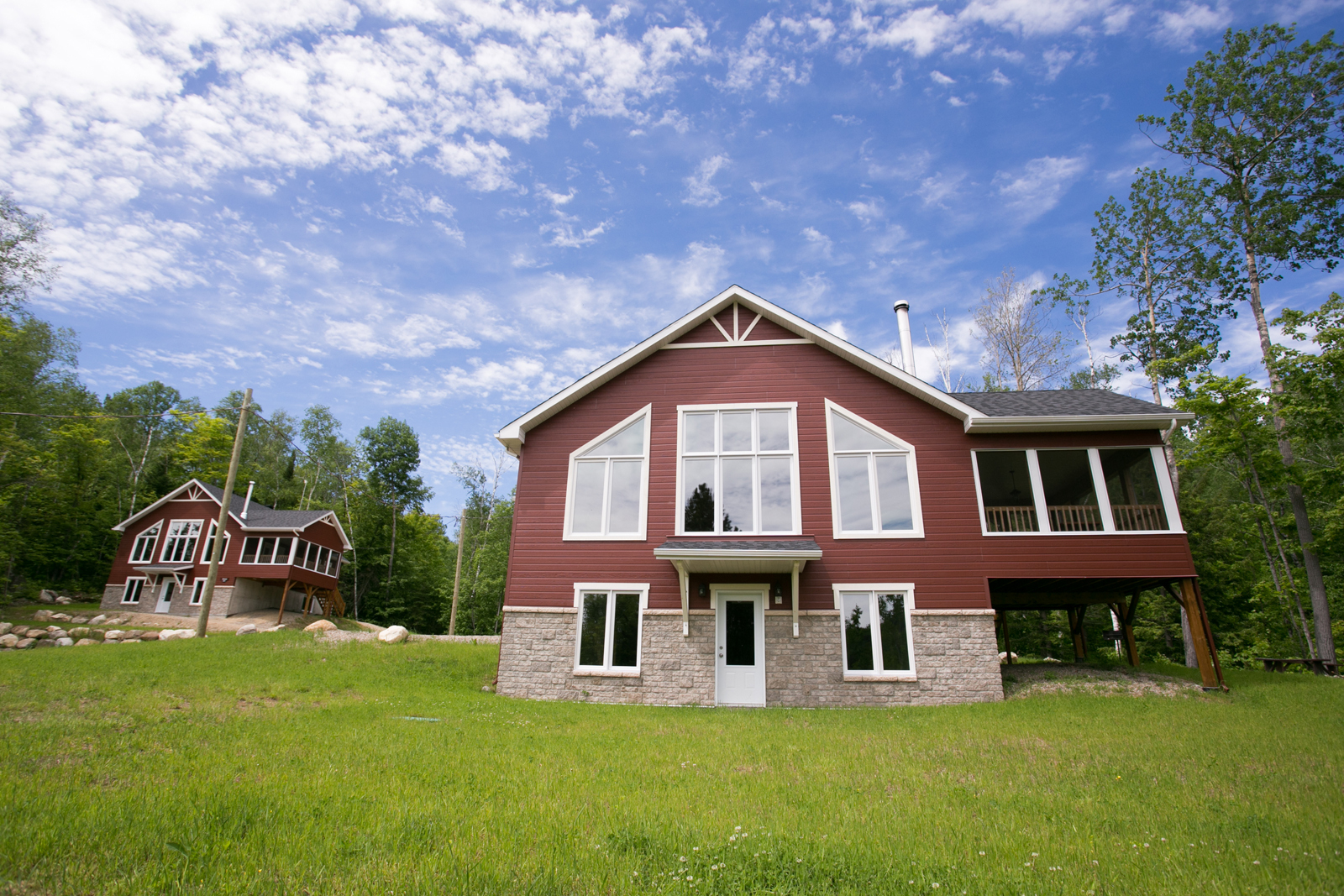 Chalet rouge avec grande pelouse, terrasse couverte et forêt environnante sous un ciel bleu.