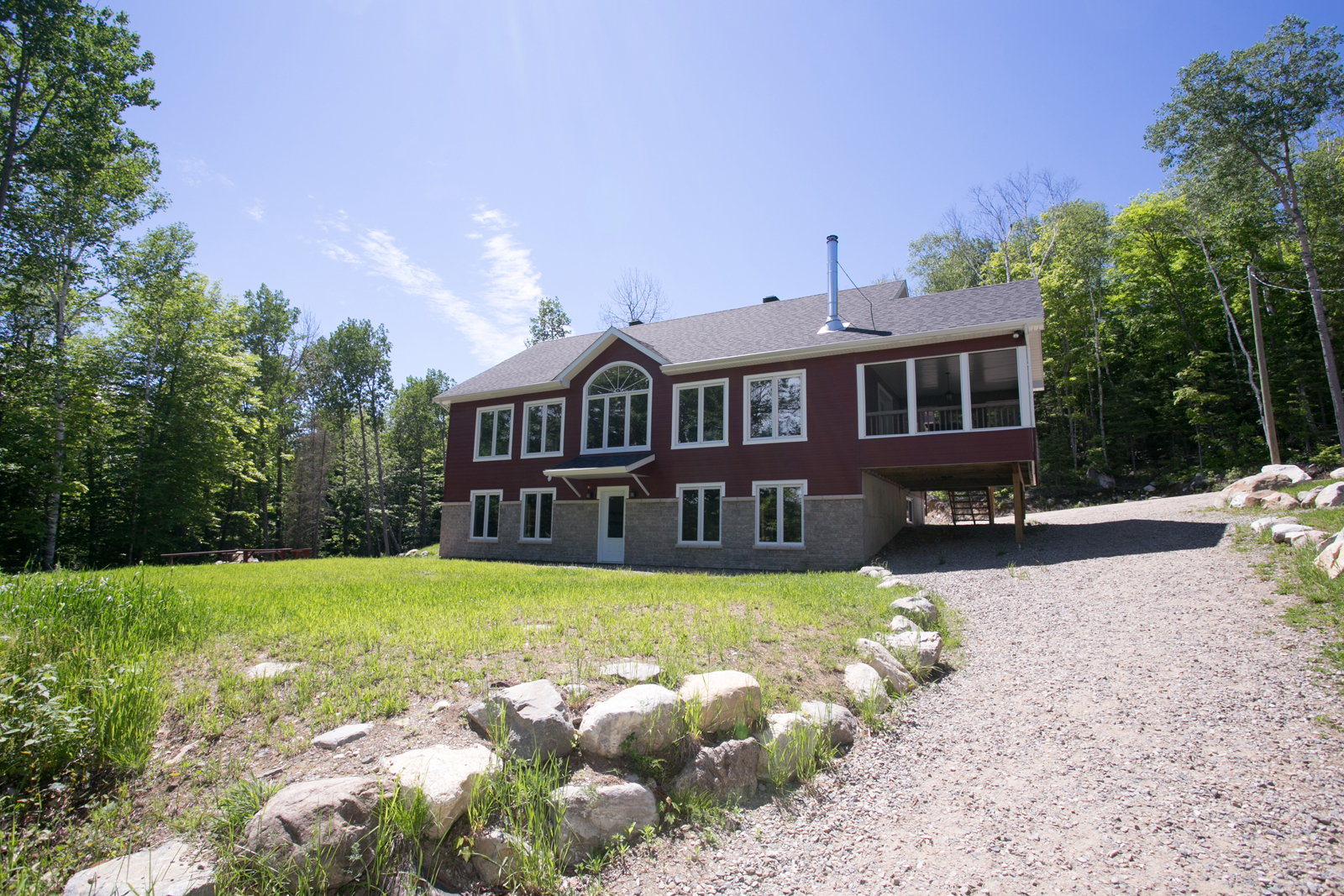 Chalet entouré d’arbres, avec grande pelouse, entrée en gravier et terrasse surélevée.