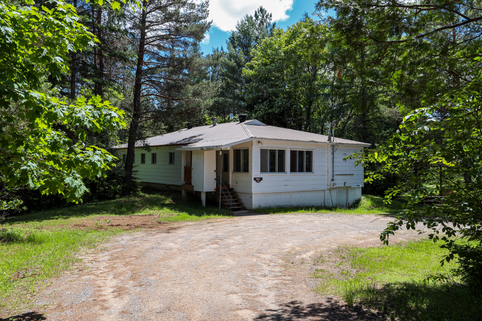 Chalet blanc entouré d’arbres, avec entrée accessible par une allée en gravier.