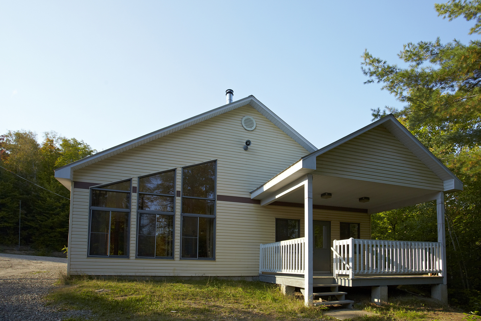 Chalet avec grande fenestration, terrasse couverte et entouré d’arbres, sous un ciel bleu.
