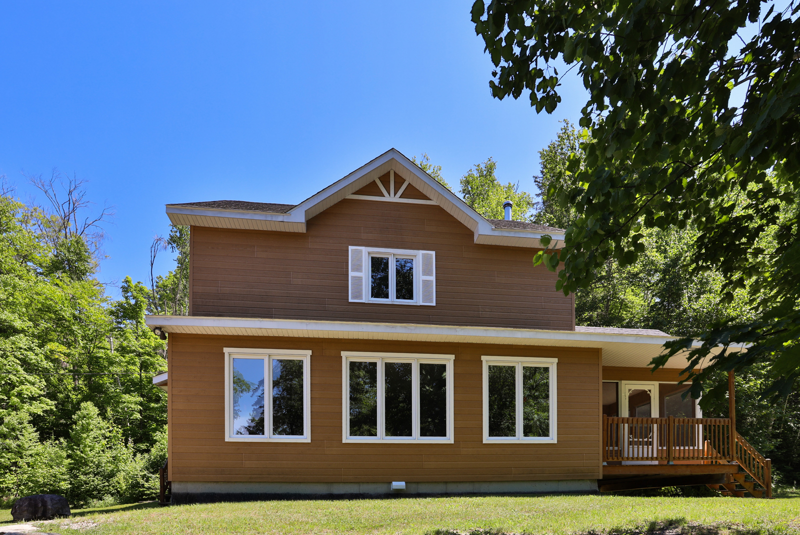 Chalet entouré d’arbres et baigné de soleil, avec terrasse et grandes fenêtres.