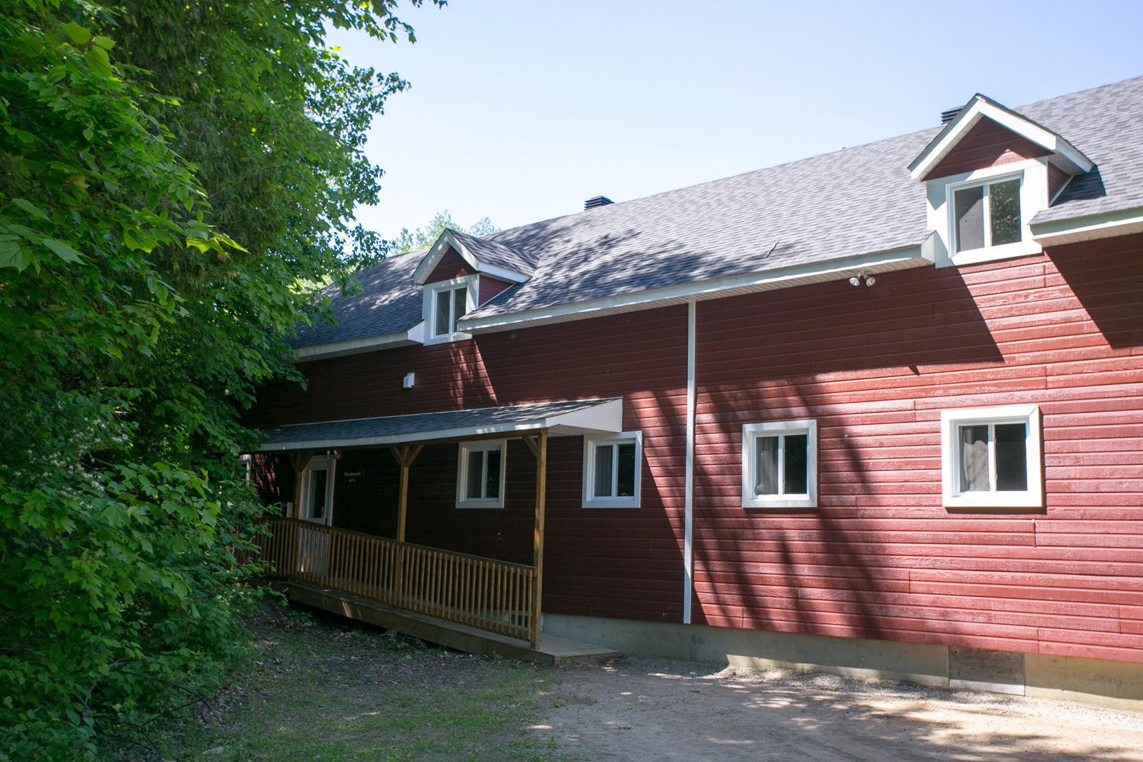 Chalet rouge entouré d’arbres, avec galerie couverte et fenêtres.