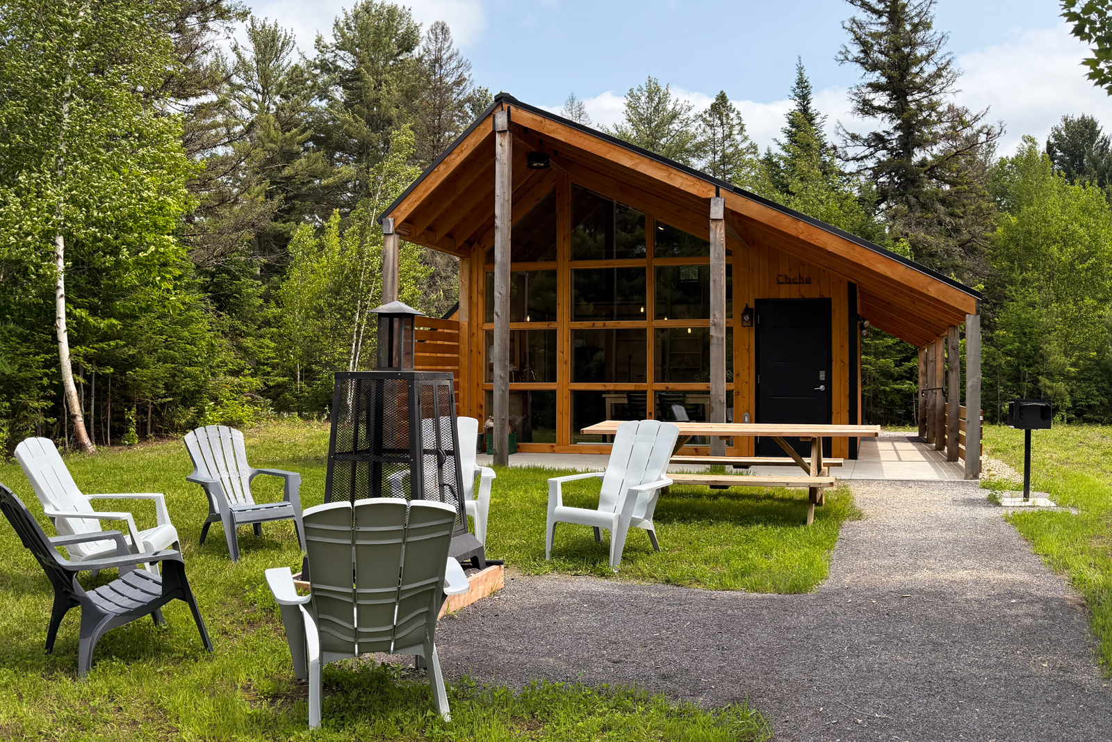 Mini-chalet en bois avec grande fenestration, terrasse, foyer extérieur et mobilier de jardin en pleine forêt.
