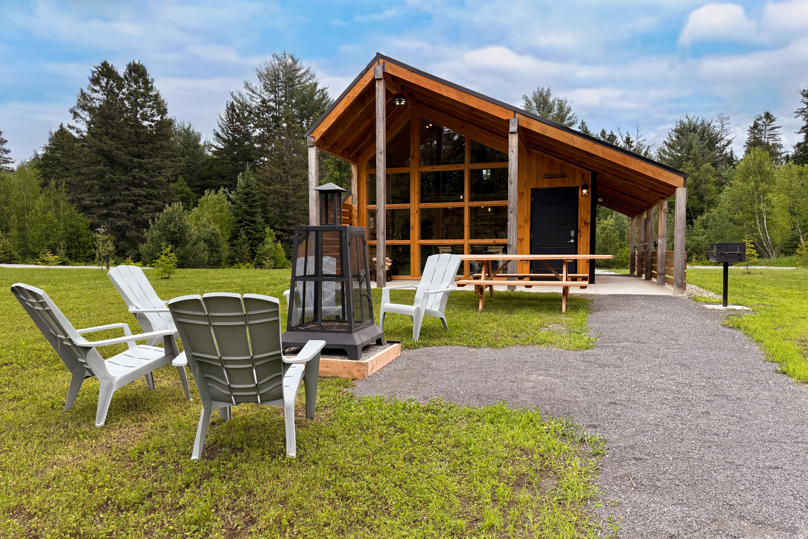 Mini-chalet en bois avec terrasse, chaises Adirondack, foyer extérieur et sentier bordé d’arbres.