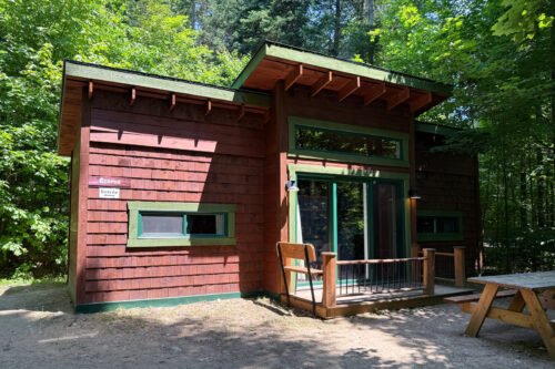 Petit cabane en bois, niché dans la forêt, avec terrasse, portes vitrées et table de pique-nique.