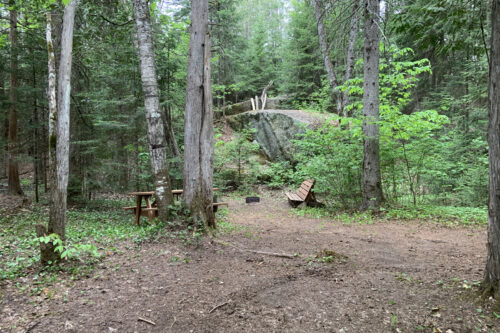 Aire de camping aménagée en forêt, avec table de pique-nique, banc et rochers.