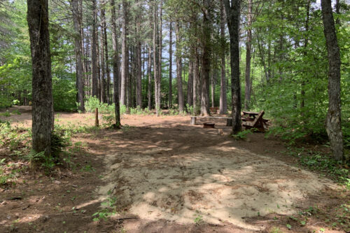 Site de camping ombragé avec tables de pique-nique, banc et aire de feu.
