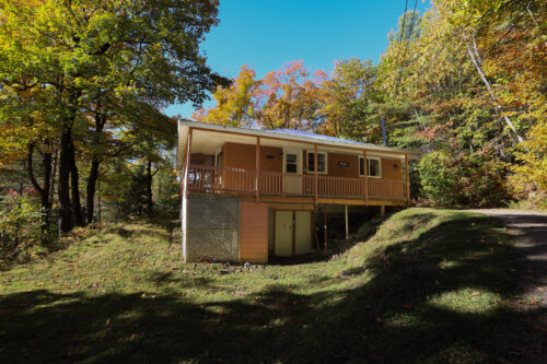 Chalet entouré d’arbres aux couleurs automnales, avec grande terrasse et sentier boisé.