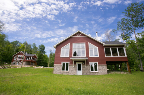 Chalet rouge avec grande pelouse, terrasse couverte et forêt environnante sous un ciel bleu.