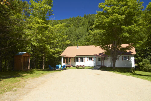 Chalet entouré d’arbres, avec aire de stationnement et mobilier extérieur sous un ciel bleu.