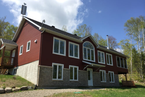 Grand chalet rouge de Chalets Lanaudière, avec grandes fenêtres, terrasse et terrain boisé sous un ciel bleu.
