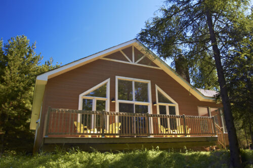 Chalet entouré d’arbres, avec grande terrasse ensoleillée et fenêtres donnant sur la nature.