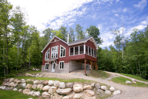 Chalet rouge entouré d’arbres, avec terrasse couverte et stationnement sous un ciel bleu.