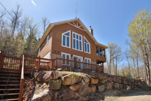Chalet entouré d’arbres, avec grande terrasse et escalier sur un terrain rocheux.