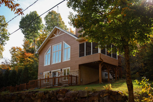 Chalet en bois entouré d’arbres, avec grande terrasse, balcon couvert et terrain rocheux aux couleurs automnales chez Chalets Lanaudière.