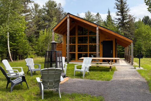 Mini-chalet en bois avec grande fenestration, terrasse, foyer extérieur et mobilier de jardin en pleine forêt.