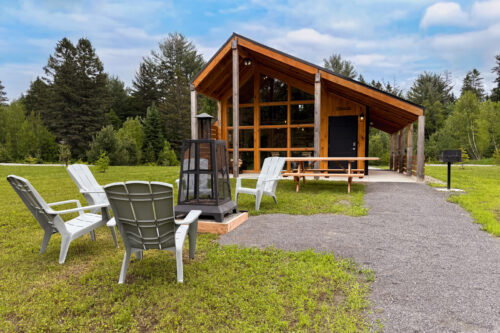 Mini-chalet en bois avec terrasse, chaises Adirondack, foyer extérieur et sentier bordé d’arbres.