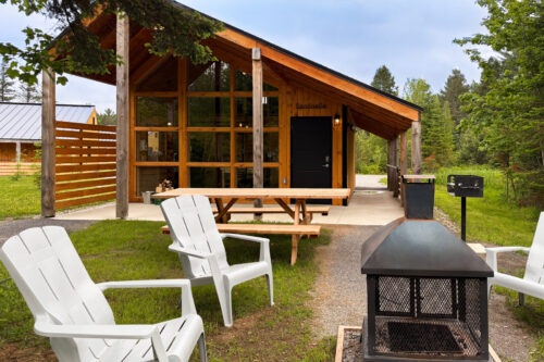 Mini-chalet en bois avec terrasse, table de pique-nique, fauteuils Adirondack et foyer extérieur dans un cadre boisé.